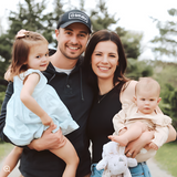 A heartwarming family portrait outdoors with co-founder Lauren. A smiling couple, with the father wearing a casual baseball cap, holds their two young children. The daughter, dressed in a pale blue dress, is cradled in her father's arm, while the mother, in a simple black top, supports their baby wearing a light beige outfit. They all share a moment of joy, capturing a snapshot of familial love and connection.