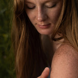 Close-up of a woman with long hair and freckles on her shoulder against a blurred green background applying tallow and honey balm.