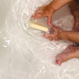 Small, delicate hands of a baby play with a bar of pale tallow soap in clear bathwater. Gentle ripples and soap suds surround the baby's hands, creating a soft and safe bathing environment. This image illustrates the gentle quality of the soap, suitable for sensitive baby skin.