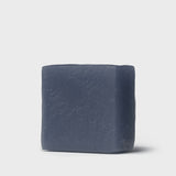 A solid cube-shaped facial soap bar with a deep, matte charcoal color sits against a light gray background. The surface texture of the soap is rustic with a slightly marbled appearance, hinting at the natural charcoal ingredient within. This soap bar is designed to detoxify and refresh the skin, as indicated by its pure and minimalist presentation.