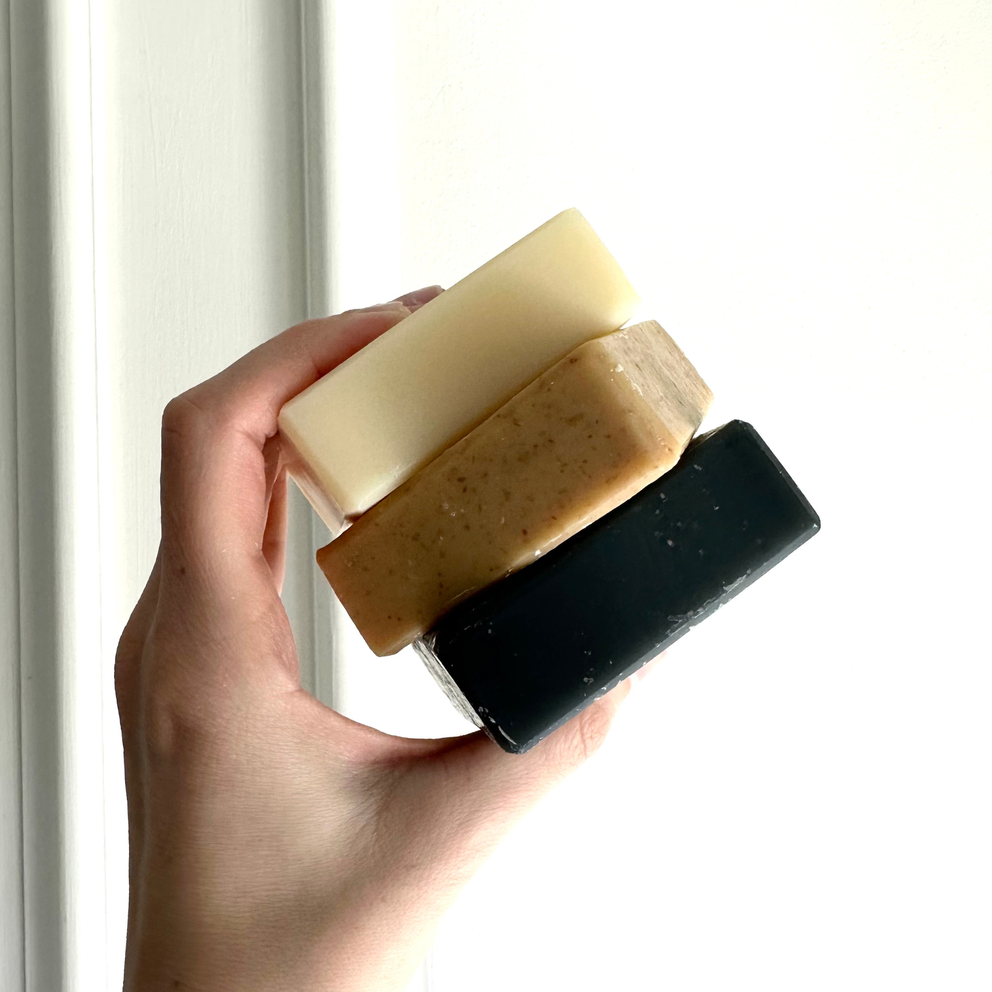 A hand holding a stack of three artisanal tallow soap bars against a light background. The bars vary in color from top to bottom: the topmost is a pale cream, the middle is a speckled tan with visible grains, and the bottom is a rich, dark charcoal. Each bar has a rustic, homemade texture, and they are positioned in a staggered arrangement, demonstrating the unique character and natural ingredients of each.