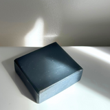 A solid black charcoal tallow soap bar rests on a white surface, bathed in natural sunlight. The angled light creates a dynamic play of shadows, accentuating the smooth, matte finish of the soap and revealing subtle textures and gentle imperfections on its surface, evoking a sense of purity and simplicity.