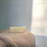 A solitary bar of Tallow Me Pretty lavender tallow soap rests on a textured beige pouf, illuminated by natural light that casts a soft glow, highlighting the soap's creamy texture and understated elegance.
