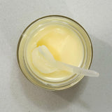 Overhead view of an open jar of Tallow & Honey Balm on a neutral background. The balm has a creamy, pale yellow color and a textured surface where a spatula has scooped out a portion, leaving a smooth trail. The spatula, with a dollop of balm, rests inside the jar, ready for application. The balm's rich texture suggests its moisturizing properties.