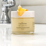 A jar of Tallow & Honey Balm is displayed against a neutral background with a dollop of golden honey artistically drizzled on top. The jar is labeled 'tallow me pretty' indicating the brand, set on a counter with a faucet in the soft-focus background, emphasizing the product's natural and hydrating ingredients.