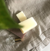 A serene composition on a natural linen fabric background featuring two artisanal Tallow Me Pretty soap bars stacked against a delicate perfume bottle with a crystal cap. The soaps exude a minimalist, handcrafted aesthetic, and a green plant leaf gently intrudes into the frame from above, adding a touch of organic vibrancy to the scene.