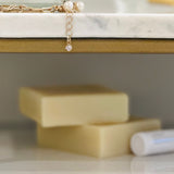A pair of neatly stacked, creamy yellow bars of soap sits on a marble countertop. In the background, a delicate pearl bracelet with a single diamond charm dangles over the edge of the shelf, adding a touch of elegance to the scene. A blurred white tube of product lies to the right, hinting at the complementary items in the skincare range.