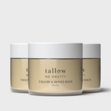 Three jars of 'Tallow Me Pretty Tallow & Honey Balm' are aligned against a white background, showcasing the product's uniform and elegant design. Each jar has a clean, white lid with the brand's signature simple typography on a golden-hued label that wraps around the container. The smooth, creamy appearance of the balm is visible through the clear glass, reflecting its natural and organic ingredients.