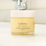 A pristine jar of Tallow & Honey Balm is placed on a white countertop, filled with a pale yellow, creamy balm. The jar's label reads 'tallow me pretty,' indicating the brand, followed by 'TALLOW & HONEY BALM' with the size 59ml/2oz. In the blurred background, a shiny bathroom faucet adds to the clean and personal care setting of the product.
