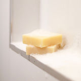 Three stacked bars of tallow soap rest on a shower ledge, immersed in frothy bubbles against a white tiled background. The soaps exude a creamy, pale yellow hue, indicating their natural tallow composition. This serene setting emphasizes the soap's use in a peaceful, cleansing routine.