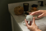 Person holding a jar of 'tallow' cream on a bathroom counter.