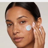 Close-up of a woman with freckles applying a dollop of light blue cream to her cheek, showing a clean, neutral background.
