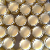 Multiple small, open jars of Tallow Balm are arranged closely together to mimic a honeycomb structure, highlighting the honey ingredient in the product. The balm inside each jar has a smooth, pale yellow appearance, reflecting the natural color of honey. The golden light shining down casts a warm hue over the array, reinforcing the natural theme and the organic essence of the balm.