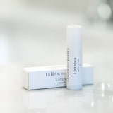 A soothing Lavender Organic Lip Balm from Tallow Me Pretty is presented upright next to its elegant white packaging, which lies horizontally. Both items are set against a soft, blurred background, emphasizing the product's natural and serene essence.
