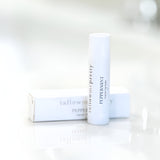A clean, minimalist presentation of a peppermint organic lip balm by 'tallow me pretty'. The open box lies horizontally next to the upright balm stick with a white cap, both on a reflective surface. The brand's name is elegantly printed in black on both the stick and the box, emphasizing the product's organic quality and inviting a sense of natural purity.